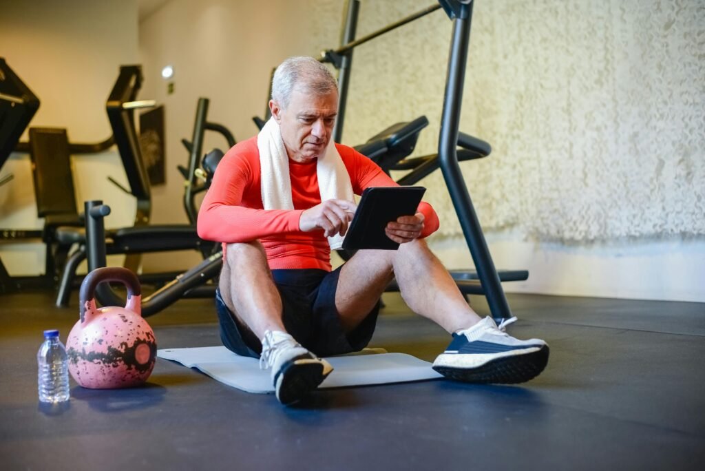 Why Do So Many Young Men Think They Have Low Testosterone? Senior man in a gym, seated on yoga mat, using a tablet for workout guidance.
